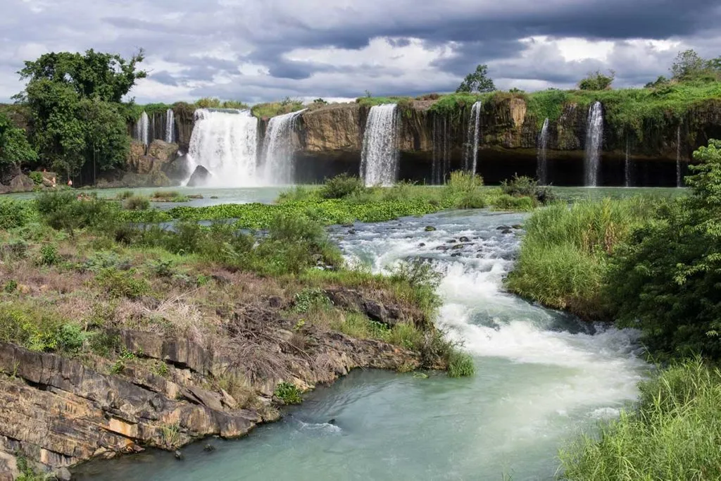 Dray Nur Waterfall in Buon Ma Thuot, Vietnam, showcasing powerful white water cascading down dark basalt cliffs amidst green vegetation.