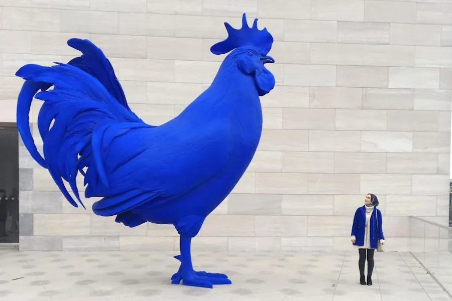 Visitor standing next to a vibrant blue rooster sculpture at the National Gallery of Art, showing the contrast between human size and the scale of the artwork.