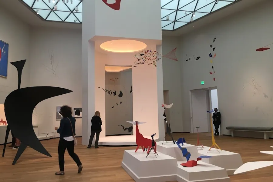 Visitors exploring the Calder Room at the National Gallery of Art, showcasing abstract and colorful mobile sculptures by Alexander Calder, hanging from the ceiling and displayed on pedestals.