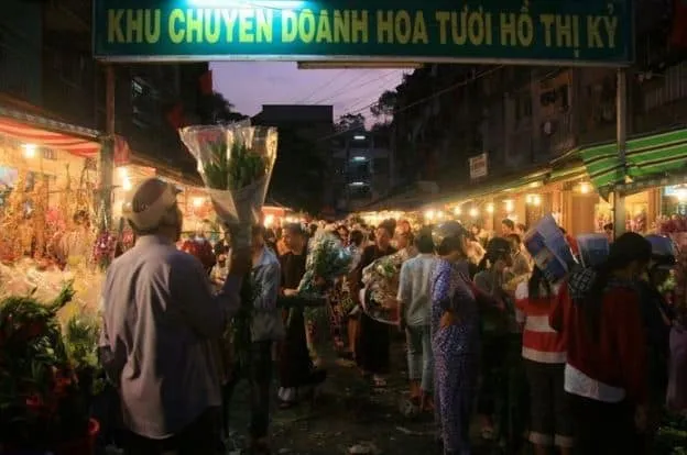 A lively and bustling scene at Ho Thi Ky Flower Market at night, with food vendors and shoppers.